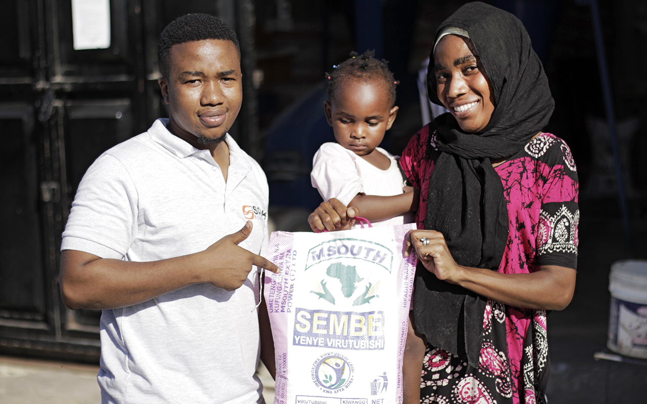 Sanku rep stands with a young mother and toddler while pointing at a bag of flour